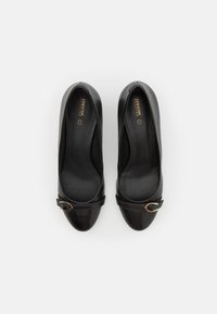Chaussures à talons hauts en cuir noir avec un bout rond, dotées d'une sangle décorative et d'une boucle métallique, semelle intérieure perforée visible.