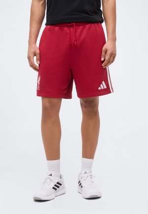 Άτομο με κόκκινα σορτς Liverpool FC με λογότυπο Adidas και λευκά παπούτσια Adidas με μαύρες ρίγες, στέκεται μπροστά σε απλό φόντο.