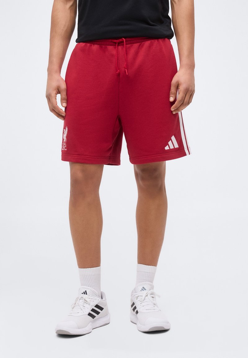 Άτομο με κόκκινα σορτς Liverpool FC με λογότυπο Adidas και λευκά παπούτσια Adidas με μαύρες ρίγες, στέκεται μπροστά σε απλό φόντο.