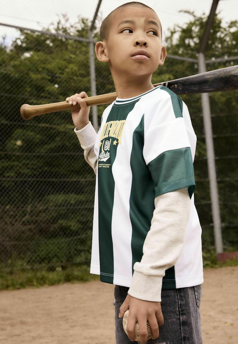 Camisa de manga corta a rayas verdes y blancas con una capa de manga larga gris debajo, sosteniendo un bate y una pelota de béisbol en un campo de béisbol.
