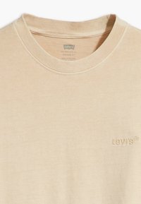 T-shirt en coton beige avec un col ras du cou classique et un logo brodé discret. Texture douce avec un design à coupe vintage.