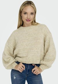 Pull en tricot beige avec une silhouette ample, ourlet et poignets à côtes, présentant des motifs texturés. Associé à un jean bleu ajusté.