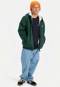 Groen vest met ritssluiting en witte nepbont voering, donkerblauw T-shirt eronder, lichtblauwe ruime spijkerbroek en oranje gebreide muts. Zwarte sneakers.