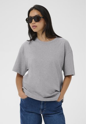 Donna che indossa una maglietta grigia oversize, jeans blu e grandi occhiali da sole neri, in piedi con le mani nelle tasche su uno sfondo uniforme.