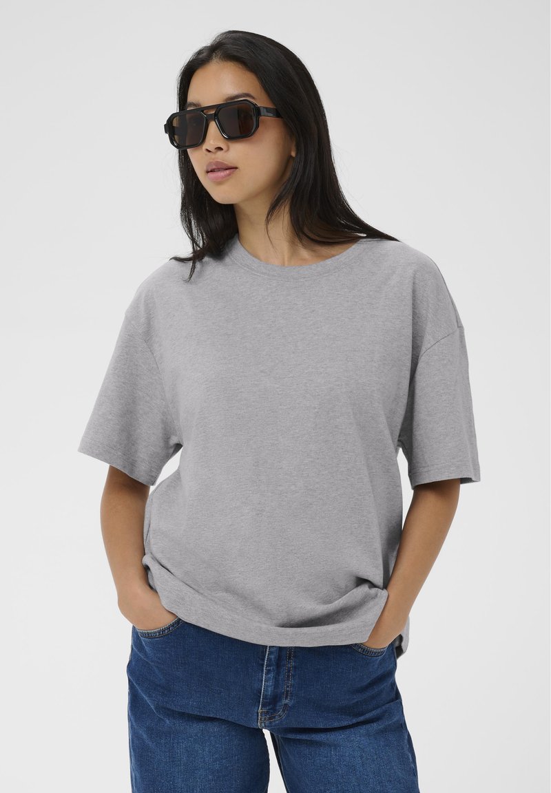 Mujer con camiseta gris de gran tamaño, vaqueros azules y gafas de sol negras grandes, de pie con las manos en los bolsillos frente a un fondo liso.