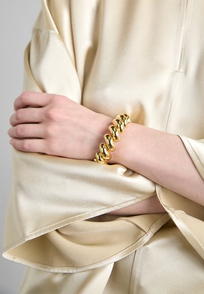 Bracelet doré présentant des maillons ovales interconnectés avec une finition lisse et polie, porté sur un poignet couvert par une manche crème.