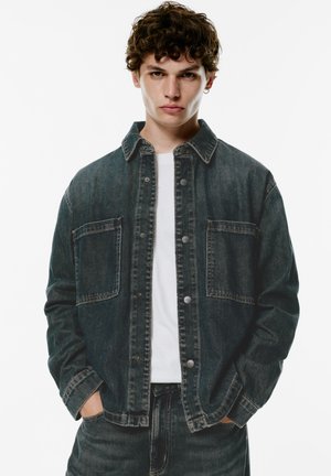 Mørkeblå denimjakke med to frontlommer, trykknaplukninger og klassisk krave. Båret over en hvid skjorte med en afslappet pasform.