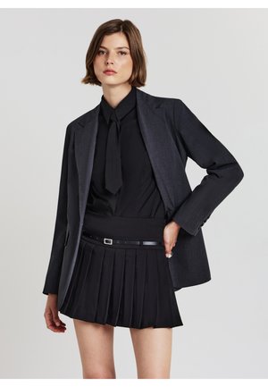 Giacca nera su misura sopra una blusa nera con cravatta e una gonna pleizzata nera. Include una cintura nera sottile con una fibbia metallica.