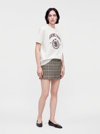 Νεαρή γυναίκα φοράει λευκό t-shirt με σχέδιο, καρό μίνι φούστα και μαύρα επίπεδα παπούτσια, στέκεται μπροστά σε ένα απλό φωτεινό φόντο.