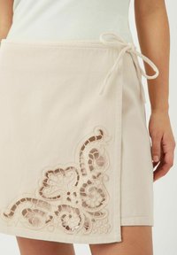Jupe portefeuille beige en coton. Présente une broderie en dentelle complexe avec un motif floral à l'ourlet et une taille à nouer. Texture lisse.