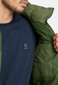 Camiseta azul marino de cuello redondo con un parche del logo de Timberland, llevada debajo de una chaqueta acolchada verde, destacando su textura suave y su apertura con cremallera.