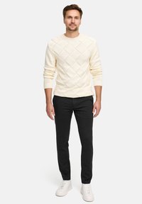Maglione a maglia crema con motivo a diamante, abbinato a pantaloni scuri a gamba affusolata e sneaker bianche, presentato su uno sfondo neutro.