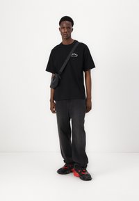 T-shirt nera oversize con grafica bianca, abbinata a jeans grigi scuri larghi e scarpe nere con dettagli rossi. Piccola borsa a tracolla nera.