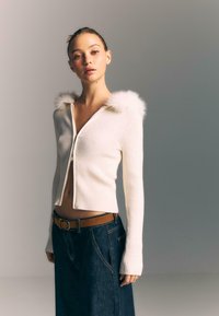 Biały kardigan z żeberkowanego materiału, zapinany na zamek, z kapturem wykończonym futrem. Noszony z ciemnoniebieską spódnicą o wysokim stanie i brązowym paskiem. Długie rękawy.