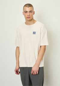 Světlé béžové oversized tričko vyrobené z bavlny, s malým modrým logem "AMERICAN VINTAGE" na levé části hrudi.