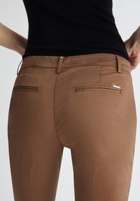 Pantaloni marroni con una texture liscia, dotati di due tasche posteriori e un design slim fit con una leggera lucentezza.