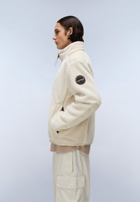 Jachetă bej din fleece cu guler înalt și fermoar, având un corp texturat, mâneci netede și o inscripție rotundă pe brațul stâng.