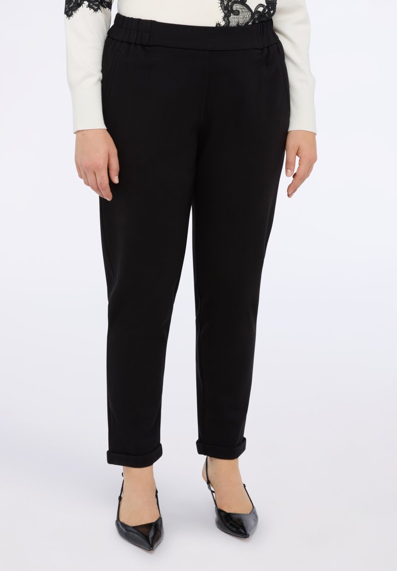 Černé kalhoty na míru s hladkou texturou, mají pružný pas a ohrnuté nohavice, doladěné s černými špičatými lodičkami.