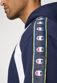 Granatowy hoodie z białymi akcentami i bocznymi paskami z powtarzającym się wzorem logo; gładka tkanina o miękkiej teksturze i sportowym kroju.