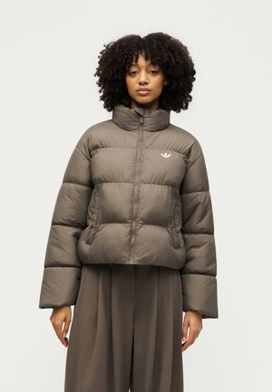 Vrouw met krullend haar draagt een grijze, gewatteerde jas met rits en hoge kraag, gecombineerd met losse bijpassende broek tegen een lichte achtergrond.