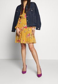 Jeansjacka med två bröstfickor över en gul blommig klänning. Lila mockahigh heels. Outfiten har livliga färger och en mjuk tygtextur.