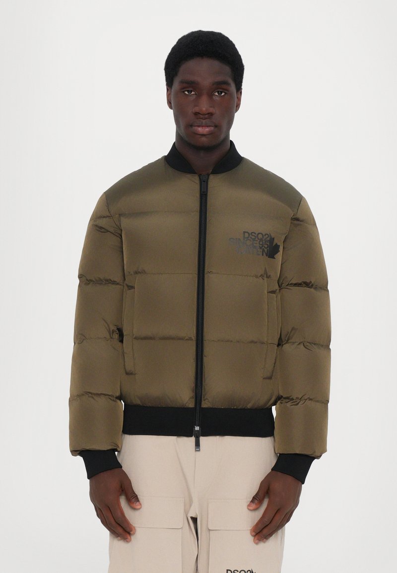 Veste bomber matelassée en olive, avec un col noir, fermeture zippée et un détail de logo. Tissu lisse et coupe structurée.