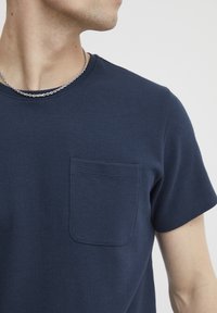 Tricou ribbenat de culoare bleumarin, cu guler rotund, mâneci scurte și un buzunar pe piept. Textură netedă, cu detalii minime. Colier din lanț argintiu.