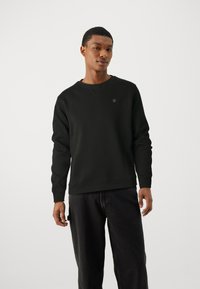 Svart sweatshirt av mjukt material, med rundad halsringning, ribbade mudd och nederkant, samt en liten logotyp på bröstet. Bärs med mörka jeans.