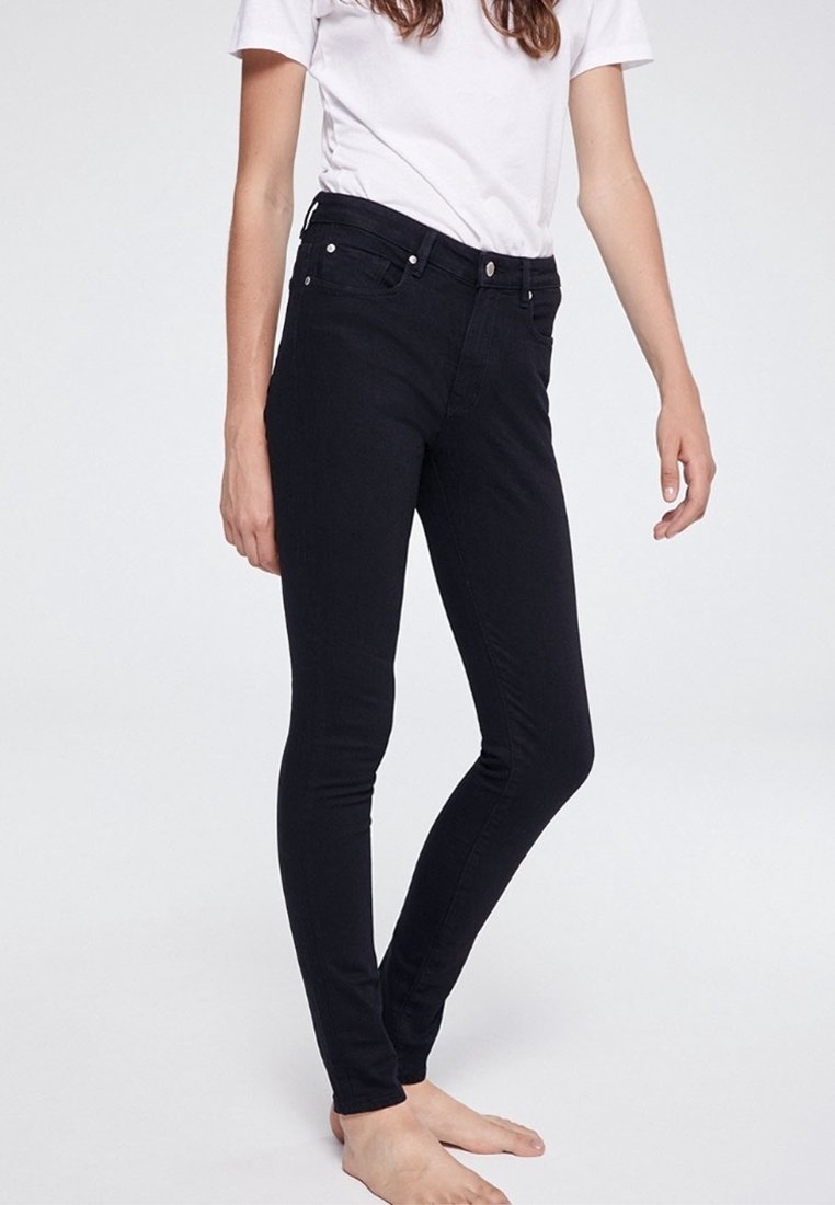Femme portant un jean noir ajusté et une chemise blanche à manches courtes, debout pieds nus devant un fond clair uni.