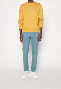Sudadera amarilla con cuello redondo y puños acanalados; pantalones rectos en color azul verdoso, de tejido ligero; zapatillas blancas con ojales de colores.