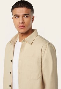 Lichtbeige overhemd met knopen en een kraag, een borstzak en een gestructureerde stof. Gedragen over een wit T-shirt; voorzien van donkere knopen.