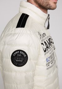Giacca a vento bianca isolata con strisce nere sulle spalle, patch rotondo del logo e testo stampato in nero e blu sul petto.