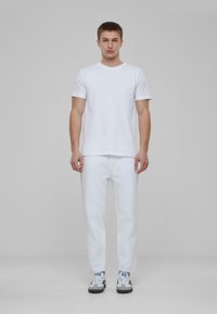 Tricou alb, blugi albi și adidași negri cu accente gri și albastre. Model în picioare, pe un fundal simplu.