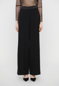 Un pantalon large noir, fabriqué en tissu lisse, présente une taille haute et des lignes épurées, porté avec des talons aiguilles noirs.