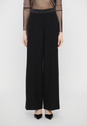 Un pantalon large noir, fabriqué en tissu lisse, présente une taille haute et des lignes épurées, porté avec des talons aiguilles noirs.