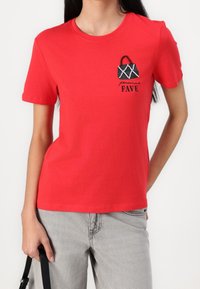 Camisa de algodón roja con mangas cortas, que presenta un gráfico de un bolso negro y la palabra "FAVE" en texto negro. Textura suave, corte casual.