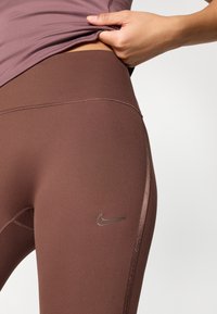 Καφέ κολάν με λεία υφή και ψηλή μέση. Διαθέτει μια διακριτική λογότυπο της Nike σε ασημί χρώμα στην αριστερή πλευρά.