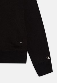 Černá mikina s měkkým povrchem, žebrovanými manžetami a malým bílým logem na zápěstí. Obsahuje boční kapsu s diskrétním švem.