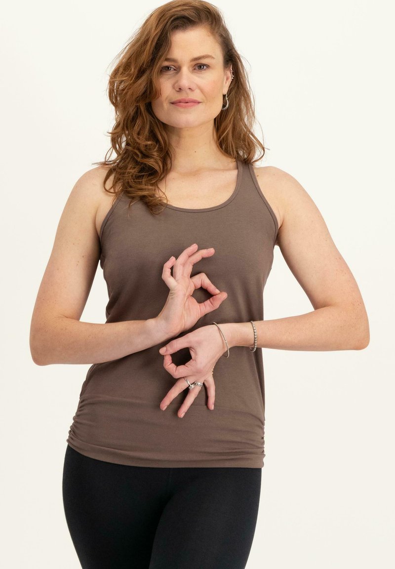 Braunes Tanktop aus weichem Stoff, mit einem figurbetonten Design und dünnen Trägern, kombiniert mit schwarzen Leggings. Hände zeigen eine Geste.
