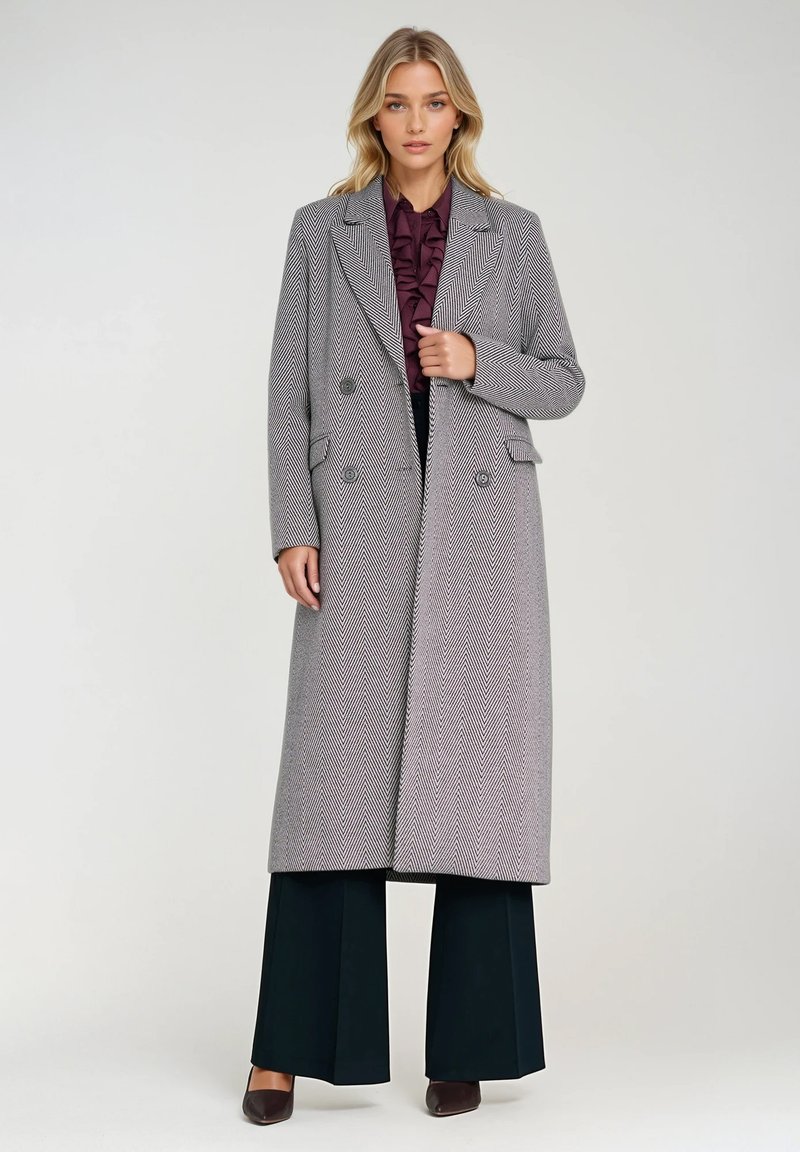 Podwójnie zapięty płaszcz w jodełkę w odcieniu szarym z oversize'owymi klapami, bocznymi kieszeniami i dopasowanym krojem, nałożony na ciemne szerokie spodnie.