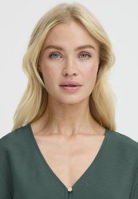 Blond naine siniste silmadega, seljas tume roheline V-kaelusega pluus, millel on väike kuldne nööp, poseerib lihtsa valge tausta ees.