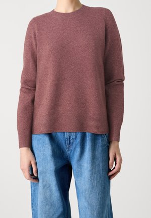 Personne portant un pull mauve à manches longues et un jean bleu ample, debout devant un fond uni.