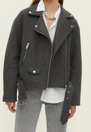 Personne portant une veste motard en laine gris foncé avec des fermetures éclair argentées et une ceinture, une chemise blanche à col, un pantalon gris et un collier torsadé en argent.