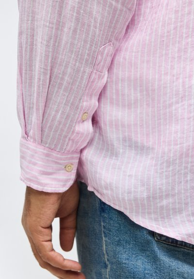 Χέρι άνδρα με πουκάμισο μακρυμάνικο ροζ και λευκές ρίγες δίπλα σε μπλε τζιν τζιν παντελόνι πάνω σε απλό φόντο.