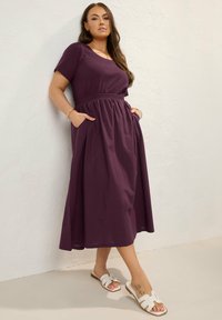Violetti, lyhythihainen mekko, jossa on sovitettu yläosa ja joustava vyötärö. Mekossa on sivutaskut ja laskostuva helma, ja se on yhdistetty valkoisiin liukuaviksi kenkiin.