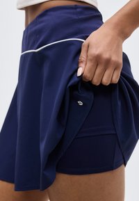 Main soulevant une jupe de sport bleu marine pour révéler un short intégré avec un petit logo blanc, montrant les détails des vêtements de sport sur la cuisse.