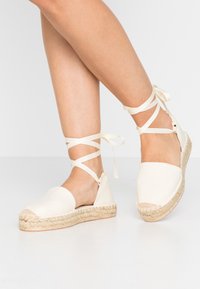 Espadrille beige avec une tige en toile, un bout rond et une semelle en corde de jute. Présente des lacets en ruban enroulés autour de la cheville pour un maintien sécurisé.