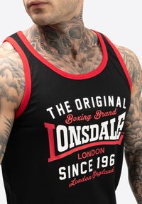 Top negru fără mâneci cu margini roșii, având un text alb și roșu îndrăzneț, "Lonsdale," și branding legat de box. Expune pielea tatuată de pe gât.