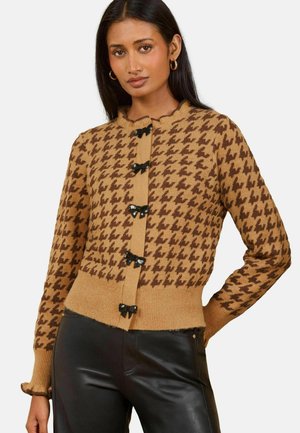 Frau trägt braune Strickjacke mit Hahnentrittmuster und schwarzen Schleifenverschlüssen sowie schwarze Lederhose, posiert mit einer Hand in der Hüfte.