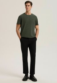 Oliwkowa koszulka z krótkim rękawem i okrągłym dekoltem, zestawiona z czarnymi spodniami na miarę i czarnymi sznurowanymi butami, o luźnym kroju.